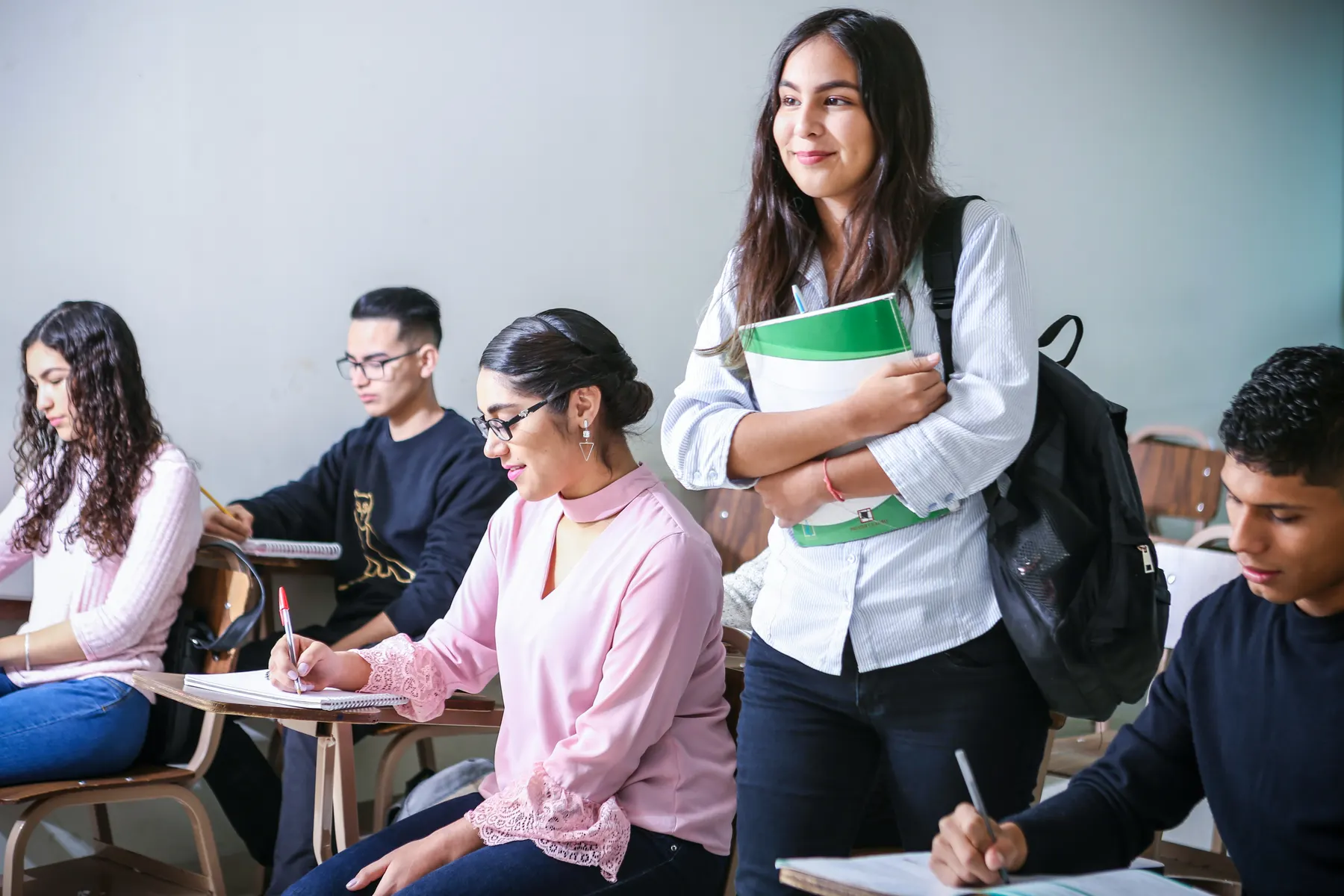 Students working at desks in a classroom while a classmate stands holding a notebook and smiling