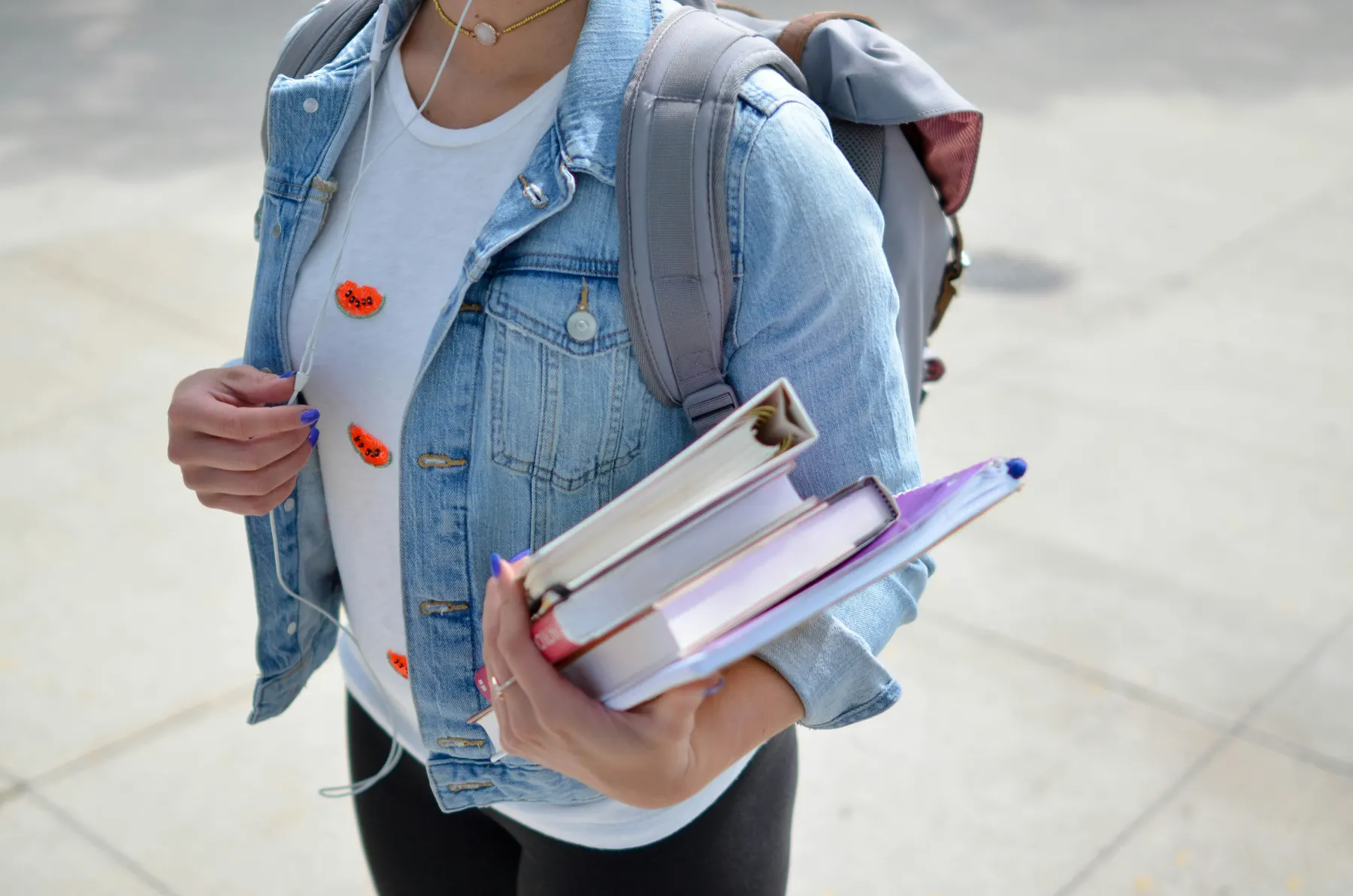A student in a denim jacket carrying a backpack and a stack of books across campus