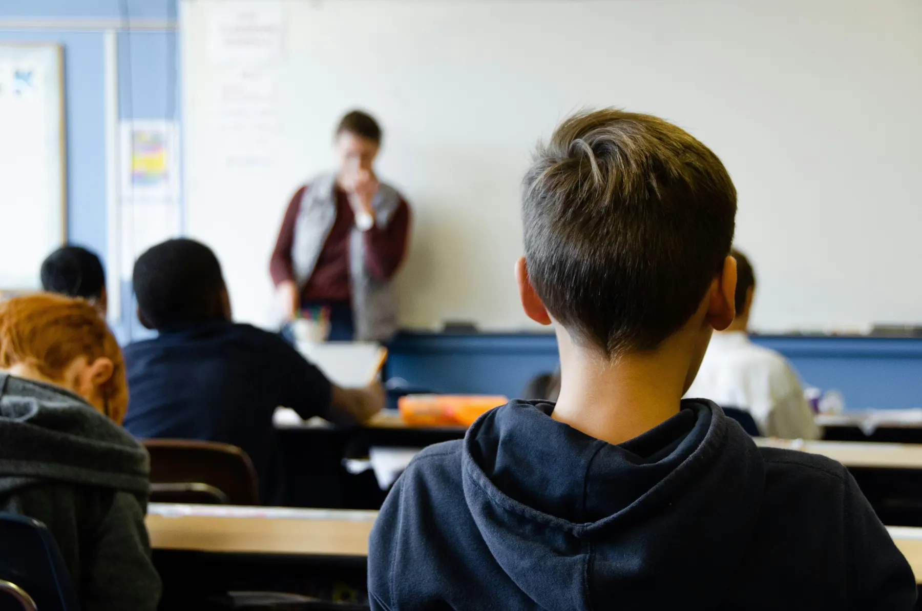 A student seen from behind in a classroom, facing a teacher at the whiteboard