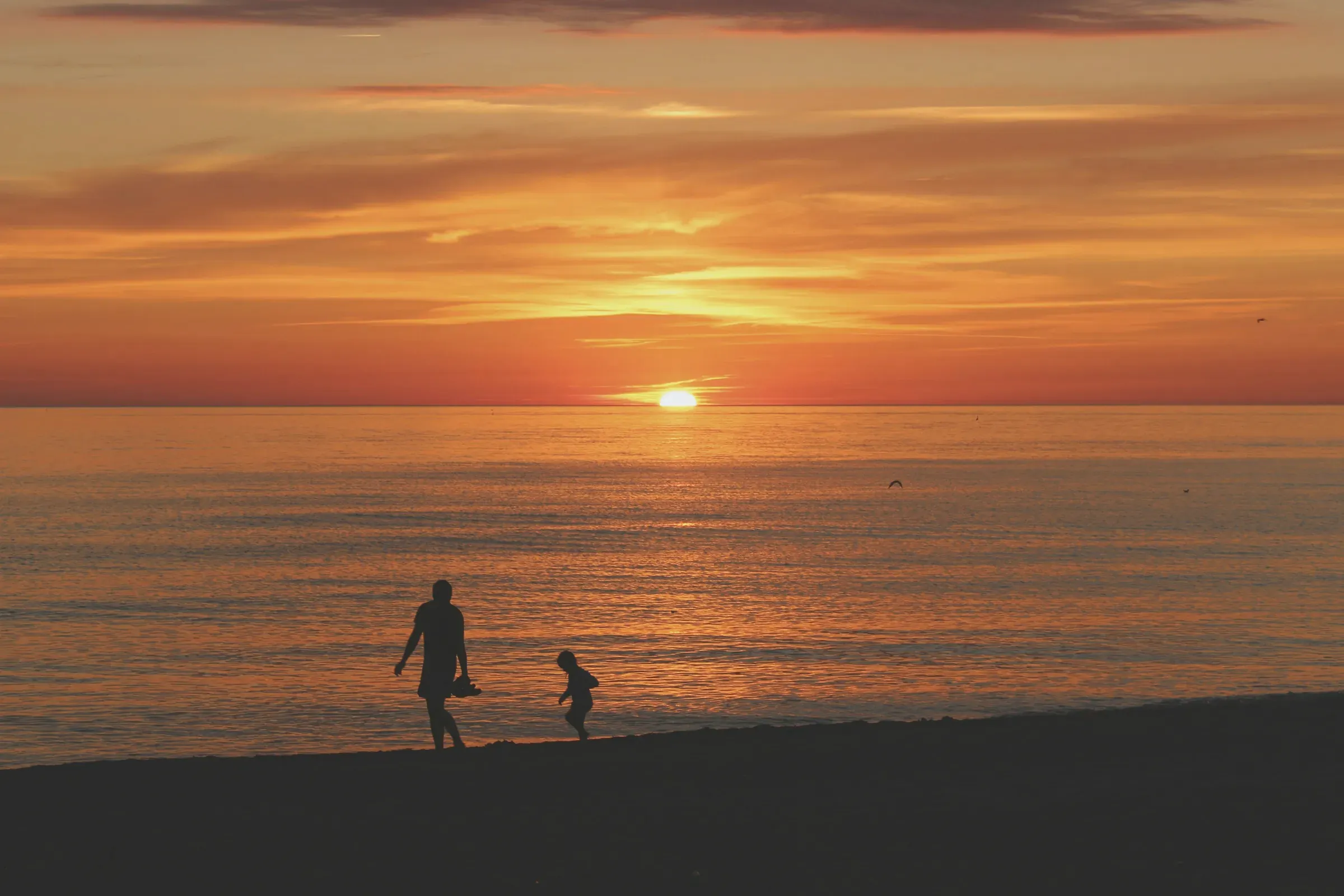 Two figures on the sand against the sun setting over the water