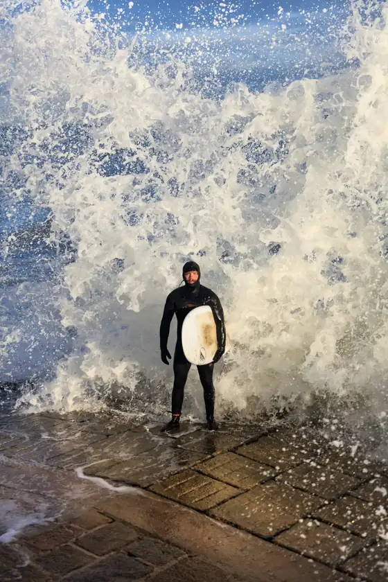 Portrait of a guide in a hooded wetsuit on the slipway, a wave breaking behind