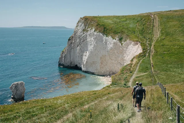 A walker on a grass path above a chalk headland and a blue cove